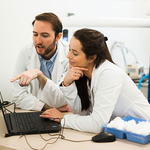 Two medical professionals, a man and a woman, are looking at a laptop screen together in a lab environment, discussing over data with engaged expressions.