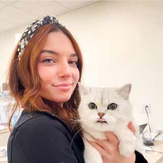 A young woman with red hair and a black headband smiles gently while holding a fluffy white cat with wide green eyes. they are in a softly lit room.